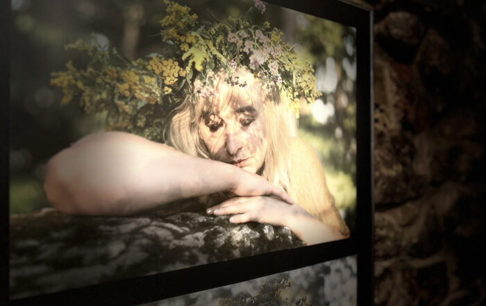 Zbliżenie na fotografię. Na fotografii portret młodej kobiety. Ma na głowie wianek. Kobieta ma zamknięte oczy i opiera twarz na rękach.