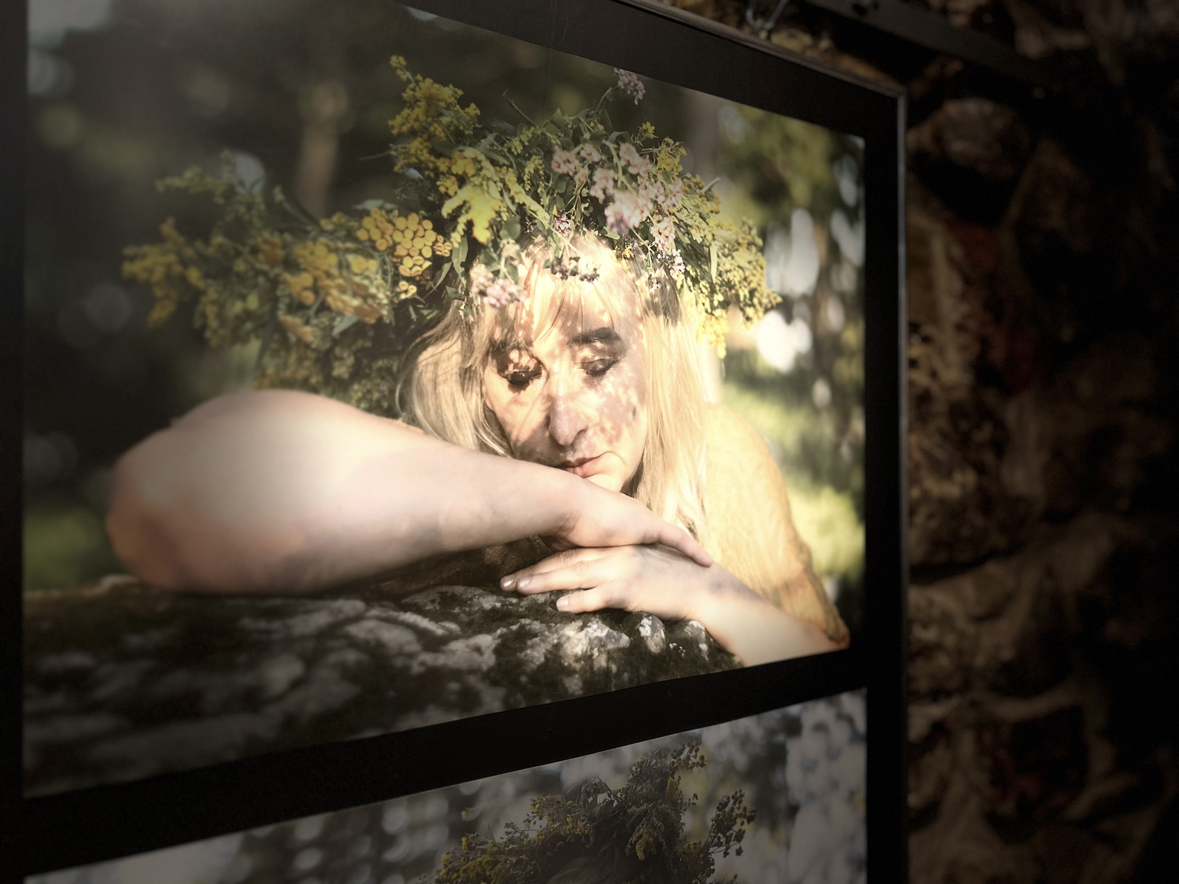 Zbliżenie na fotografię. Na fotografii portret młodej kobiety. Ma na głowie wianek. Kobieta ma zamknięte oczy i opiera twarz na rękach.