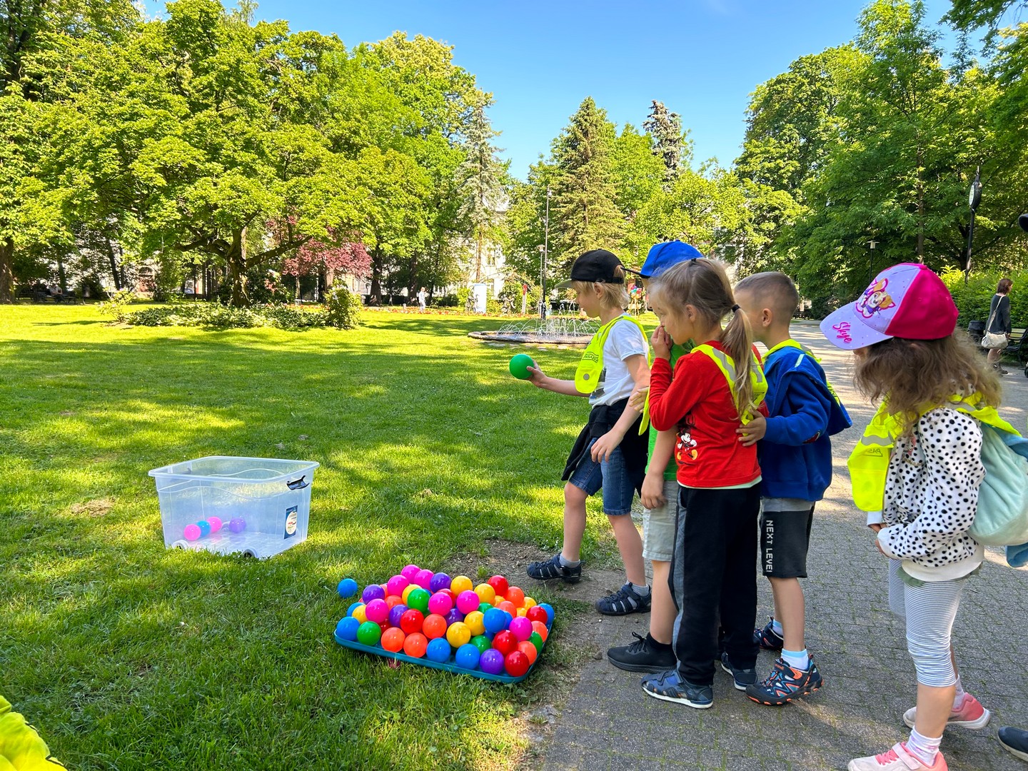 dzien_dziecka_planty05 Park miejski, słoneczna pogoda, duży trawnik, w głębi zielone drzewa liściaste. Po prawej stoi grupa dzieci, obok, na ziemi małe, kolorowe piłeczki. Jeden z chłopców trzyma w ręku zieloną piłeczkę, celuje do położonego na trawniku pudełka.