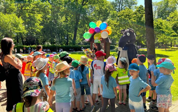 Park miejski, słoneczna pogoda, w tle dużo zielonych drzew liściastych. Blisko stoi grupa dzieci w niebieskich koszulkach i kolorowych czapeczkach. Słuchają kobiety czytającej książkę, obok niej stoi duża, bajkowa postać Wilczka.