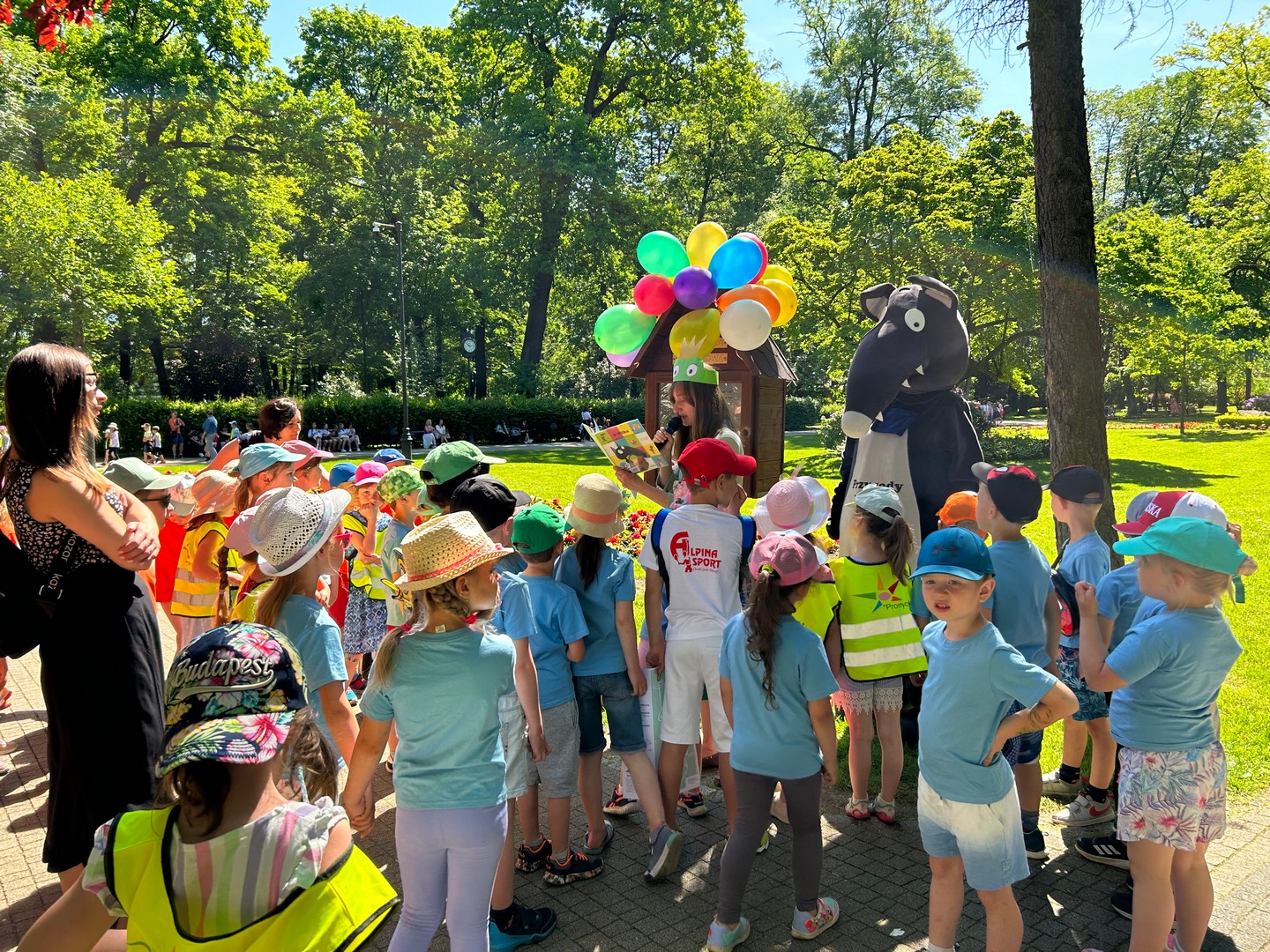Park miejski, słoneczna pogoda, w tle dużo zielonych drzew liściastych. Blisko stoi grupa dzieci w niebieskich koszulkach i kolorowych czapeczkach. Słuchają kobiety czytającej książkę, obok niej stoi duża, bajkowa postać Wilczka.