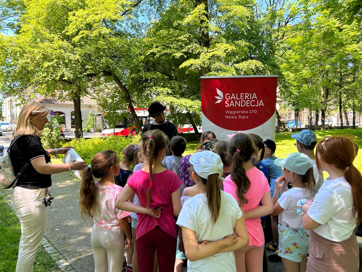dzien_dziecka_planty17 Park miejski, w tle zielone drzewa liściaste. W centrum zdjęcia stoi mężczyzna w czarnym kapeluszu i pelerynie. Wokół niego grupa dzieci. Obok stoi baner z informacjami: Galeria Sandecja Węgierska 170 Nowy Sącz.