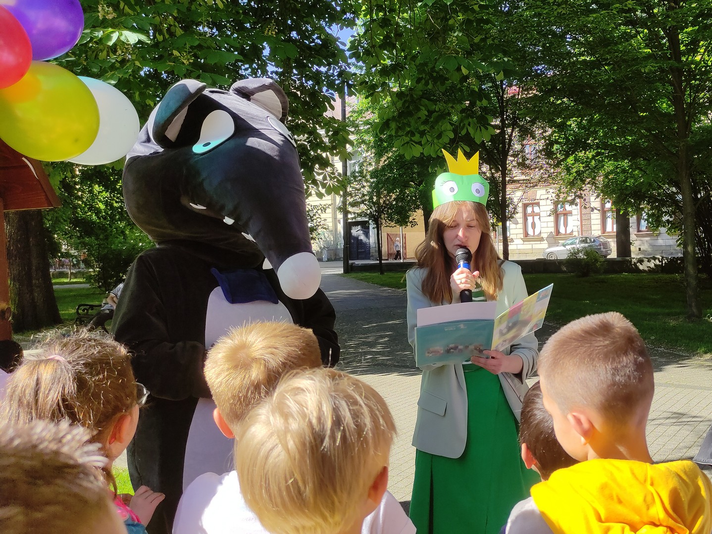 dzien_dziecka_planty19 Park miejski, w tle zielone, liściaste drzewa. Kobieta ubrana na zielono w opasce żaby w koronie. Czyta książkę, obok niej ogromna postać bajkowego Wilczka. Przed nimi kilkoro dzieci.
