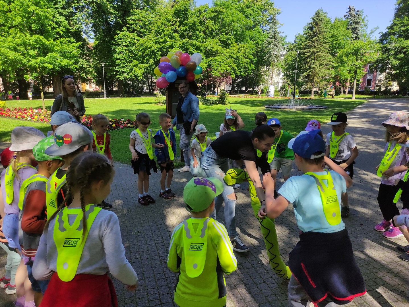 dzien_dziecka_planty22 Park miejski, słoneczna pogoda, w tle zielone drzewa liściaste. W centrum budka z wieloma kolorowymi balonami na dachu, przed budką, na alejce dzieci stoją w kręgu, w środku kręgu kobieta rozwija długiego pluszowego węża.