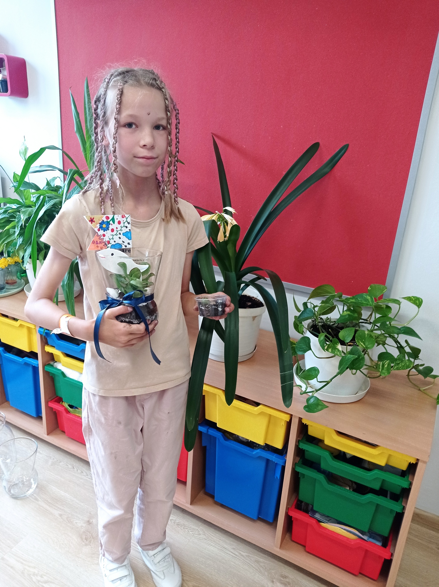 Warsztaty florystyczne_6 Dziewczynka z warkoczykami trzyma w rękach słoik i plastikowy pojemnik z posadzonymi roślinami. W tle regał z kolorowymi pojemnikami, na nim zielone rośliny.