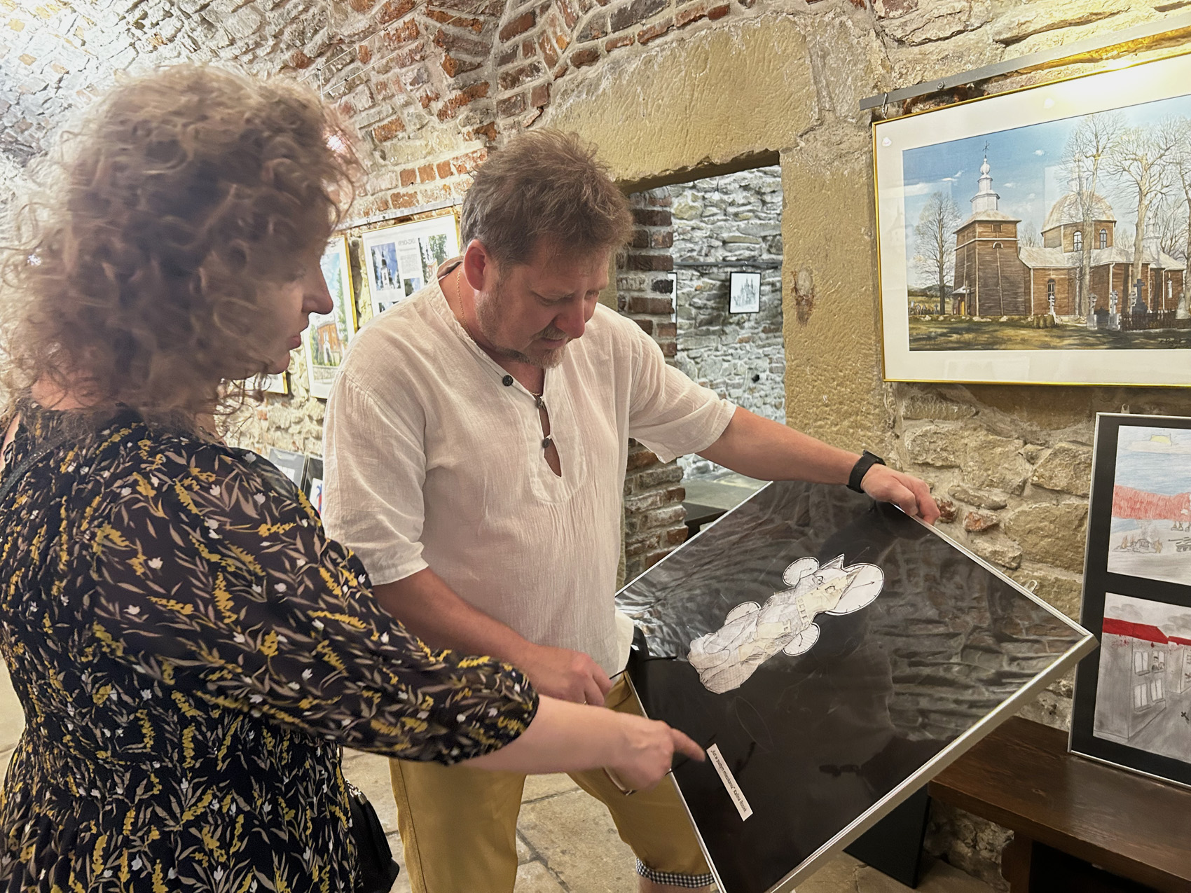 W pomieszczeniu dwójka dorosłych osób: kobieta i mężczyzna. Po lewej stronie kobieta pokazana z boku. Kobieta ma na sobie sukienkę w drobne kolorowe wzory. Obok kobiety stoi mężczyzna w białej koszulce. Mężczyzna trzyma w rękach obraz oprawiony w ramę. Kobieta i mężczyzna patrzą na obraz.