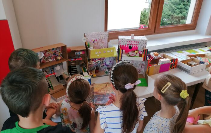 Piątka dzieci stoi przy stole, na którym są kolorowe makiety domków w kartonowych pudełkach. Dzieci oglądają je.