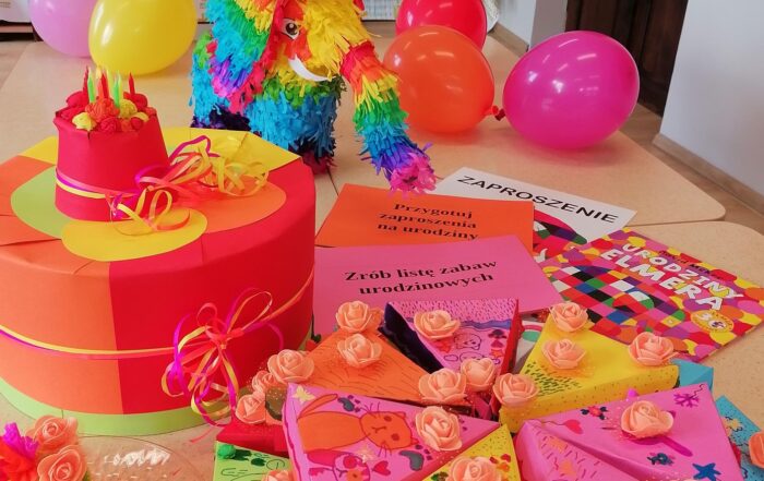 Stół z kolorową piniatą, kawałkami tortu zrobionego z kolorowego kartonu, balonami oraz kolorowymi czapeczkami urodzinowymi.