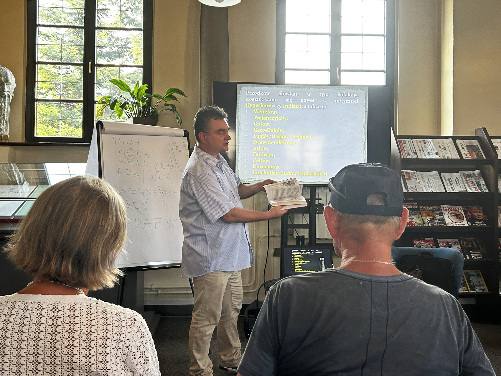 W pomieszczeniu grupa trzech osób. Kobieta i mężczyzna pokazani są od pasa w górę. Przed nimi na środku stoi mężczyzna. Mężczyzna obrócony jest bokiem, trzyma w rękach rozłożoną książkę. Obok mężczyzny po lewej stronie stoi flipchart z białymi kartkami. Za mężczyzną znajduje się ekran telewizora a obok regał z gazetami.