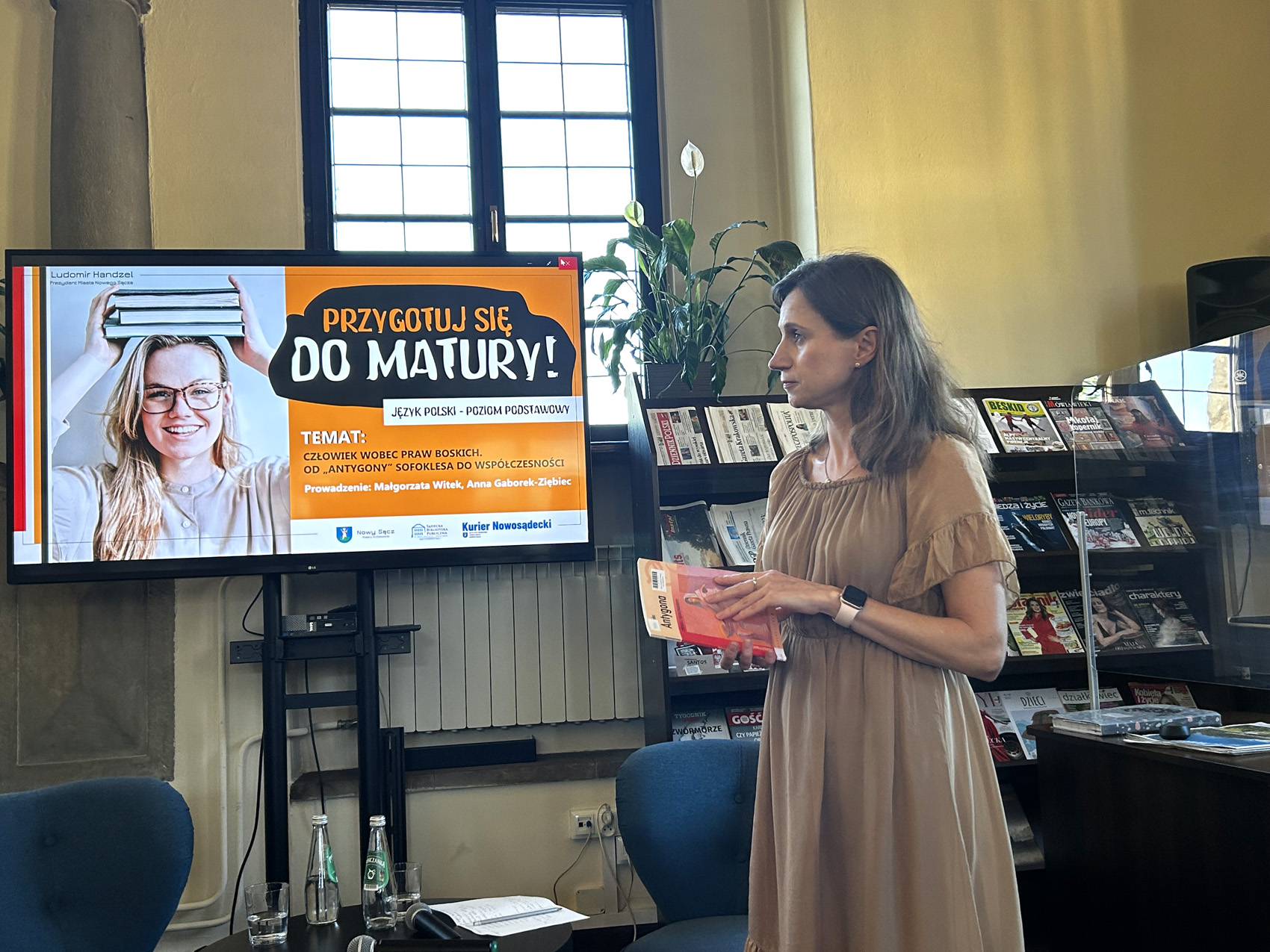 W pomieszczeniu stoi kobieta pokazana z boku, od kolan w górę. Kobieta trzyma w ręce książkę. Za nią po lewej stronie stoi duży ekran. Obok ekranu przy ścianie po prawej stronie stoi długi regał z czasopismami.