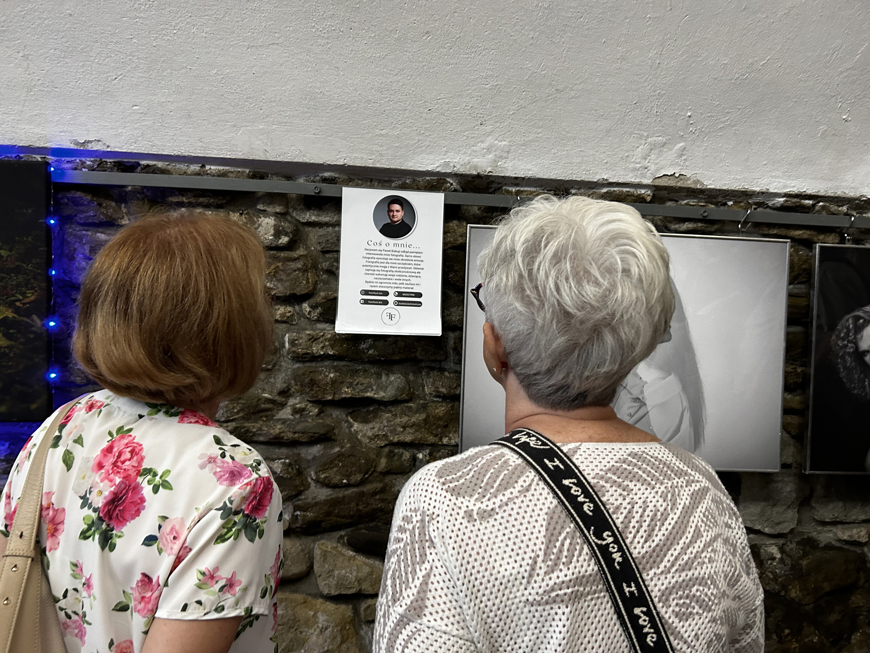 W pomieszczeniu stoją dwie kobiety, pokazane od tyłu. Przed nimi na ścianie wiszą obrazy i kartka z tekstem. Kobiety czytają tekst na kartce.