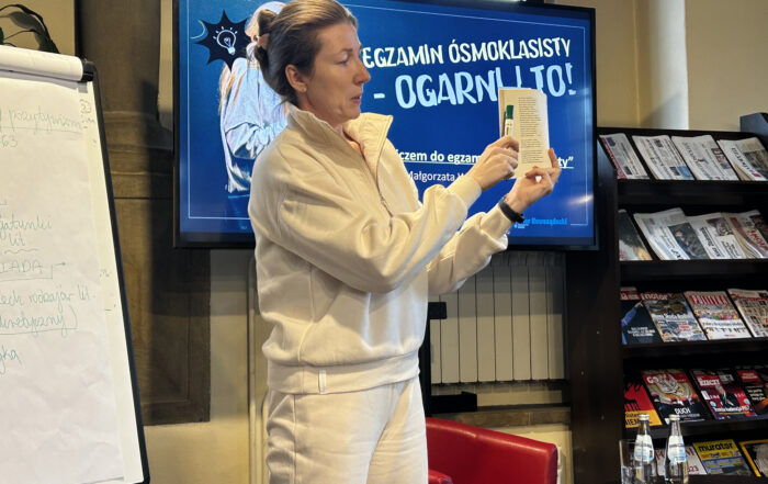 W pomieszczeniu stoi kobieta. Kobieta ubrana jest w kremowy dres. W jednej ręce trzyma otwartą książkę, w drugiej pisak. Pisakiem wskazuje na stronę książki. Obok kobiety po lewej stronie widać fragment flipcharta z kartkami. Za kobietą po prawej stronie stoi czerwony fotel. W tle duży ekran, wyświetlający grafikę.