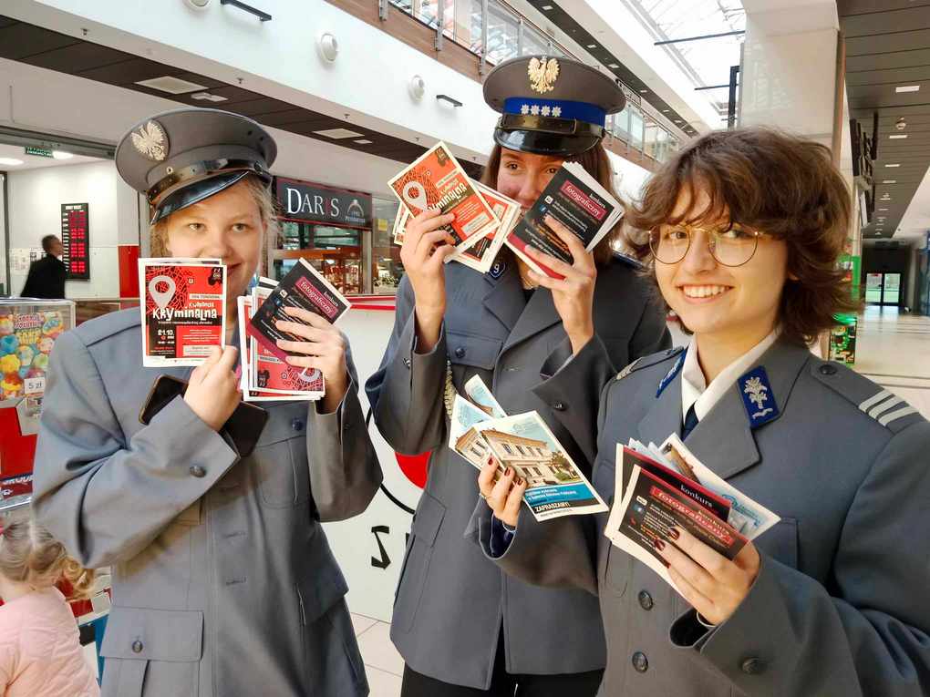 Happening policja_1 Trzy uśmiechnięte młode policjantki patrzą w obiektyw. Trzymają w rękach kolorowe ulotki. W tle wnętrze galerii handlowej.
