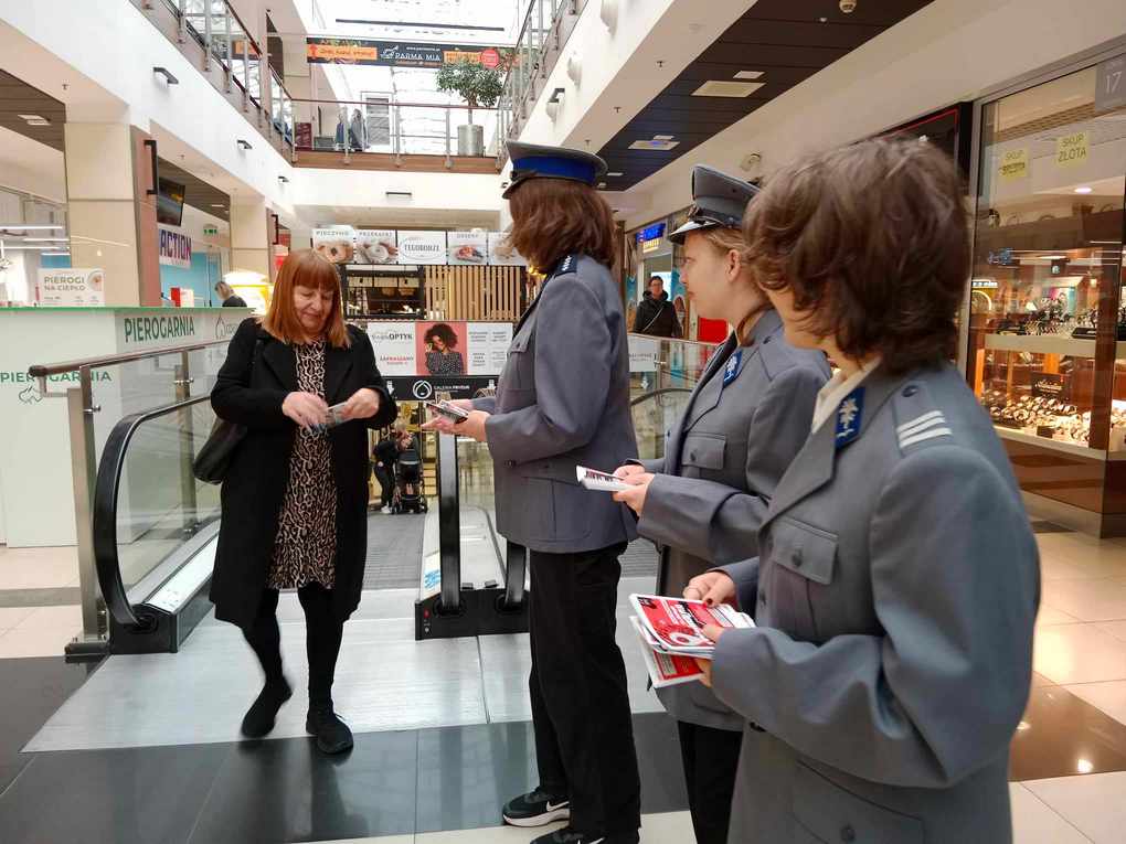 Happening policja_2 Po prawej stronie trzy policjantki stoją bokiem i patrzą w stronę kobiety, która spogląda na ulotkę. W tle galeria handlowa.