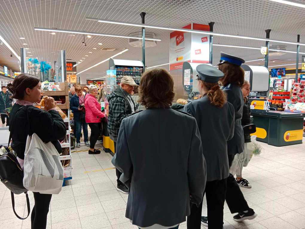 Happening policja_3 W tle wnętrze supermarketu, przy kasie kolejka ludzi. Na froncie trzy policjantki zwrócone twarzą w stronę ludzi.