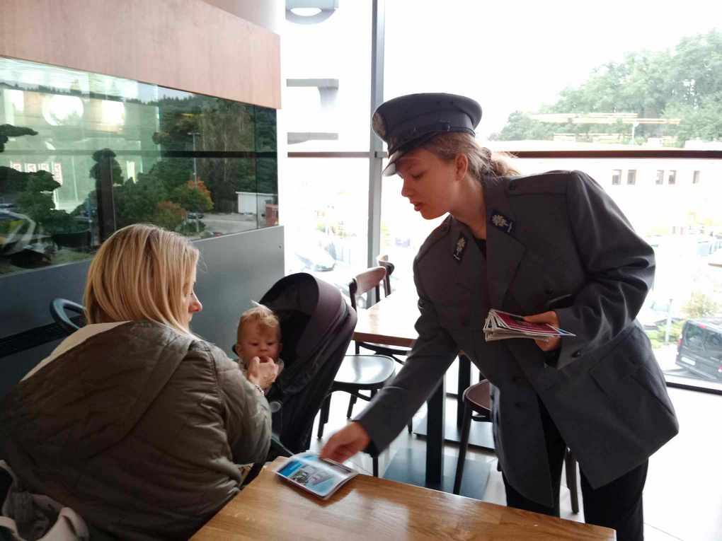 Happening policja_4 Po lewej stronie kobieta z małym dzieckiem w wózku, która siedzi przy stole. Po prawej stronie policjantka nachyla się w stronę kobiety i kładzie ulotkę na stole przed nią.