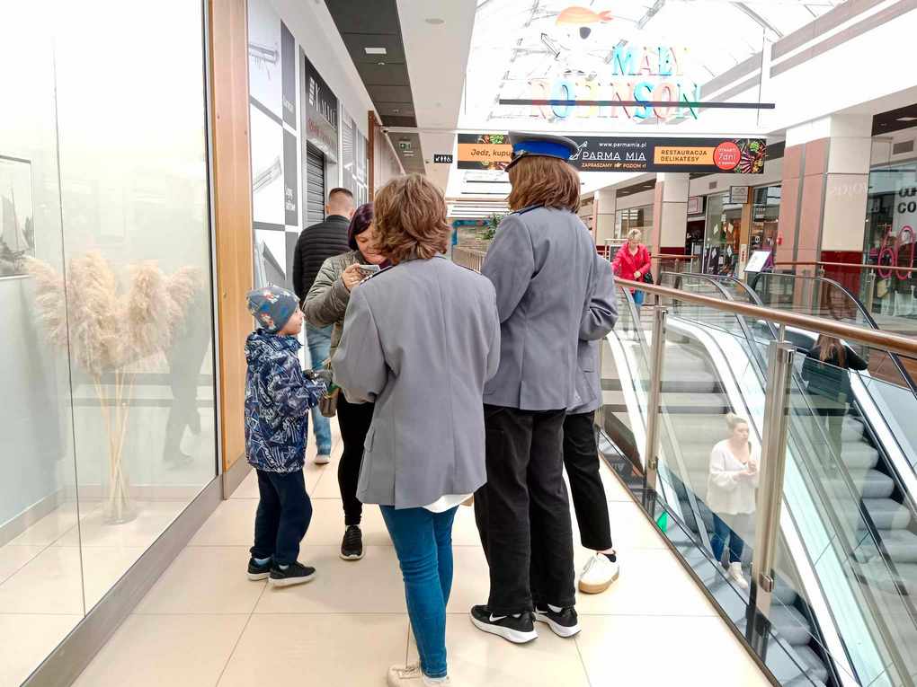 Happening policja_5 Korytach w galerii handlowej. Po prawej stronie ruchome schody. Po lewej trzy osoby w policyjnych marynarkach wręczają ulotki kobiecie z dzieckiem.