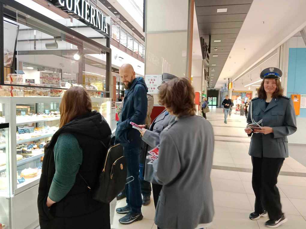Happening policja_6 Korytarz w galerii handlowej. Po lewej stronie stoisko ze słodyczami, przy którym stoją trzy osoby. Za nimi trzy osoby ubrane w policyjne marynarki z kolorowymi ulotkami w rękach.