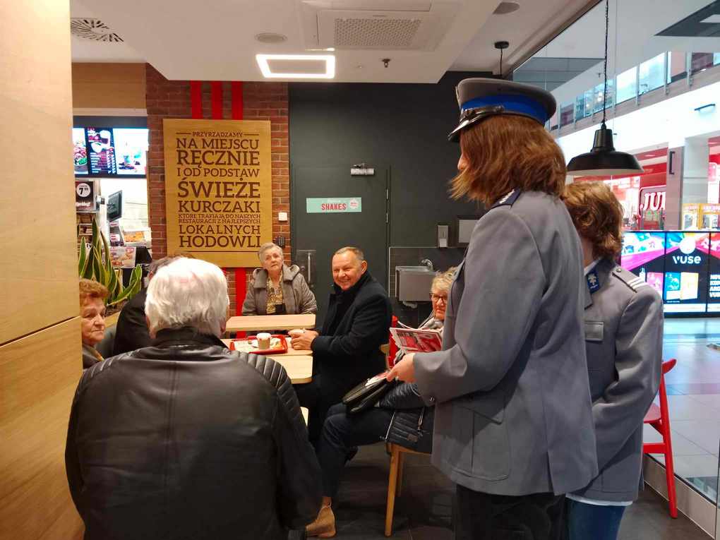 Happening policja_7 Przy stole siedzi 6 osób. Spoglądają one w stronę dwóch osób w policyjnych marynarkach, które trzymają kolorowe ulotki.