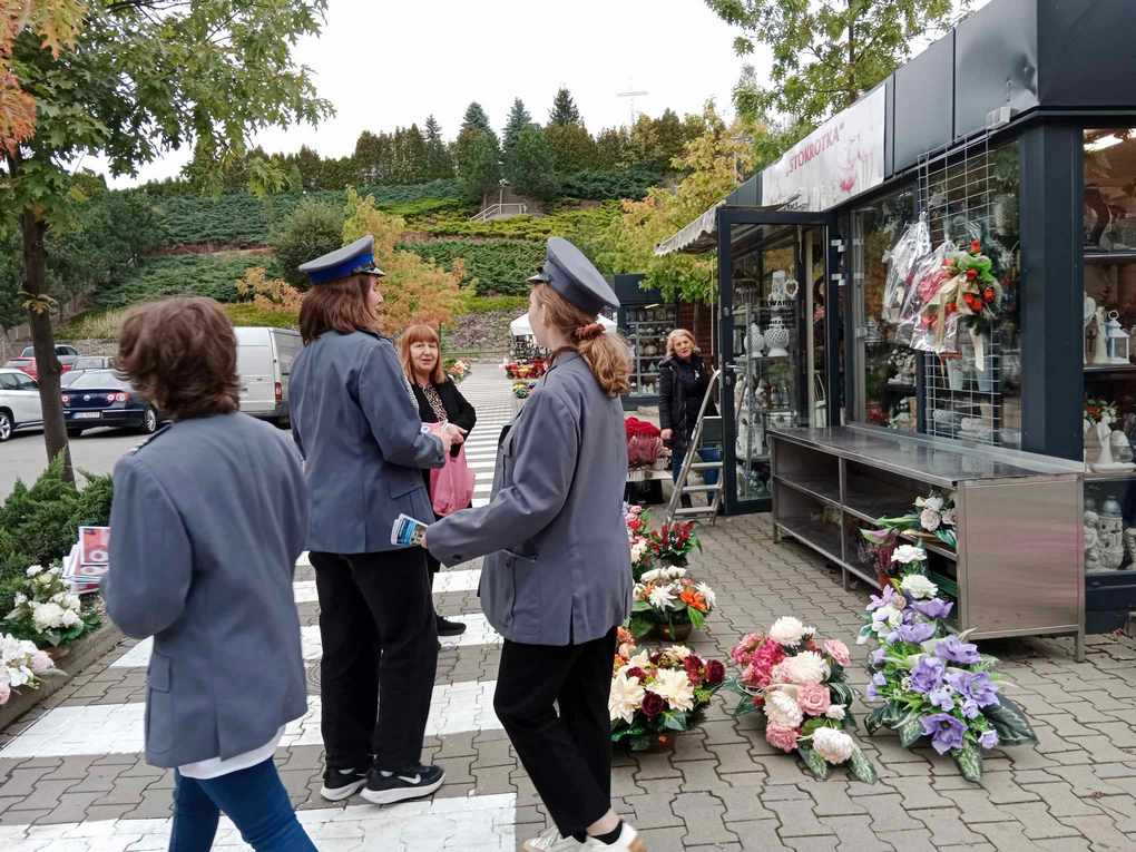 Happening policja_8 Po lewej stronie trzy osoby przebrane w policyjne marynarki trzymają w ręku kolorowe ulotki. Po prawej stronie sklep z pamiątkami. Przed sklepem wieńce z kolorowych kwiatów. W tle zielone wzgórze.