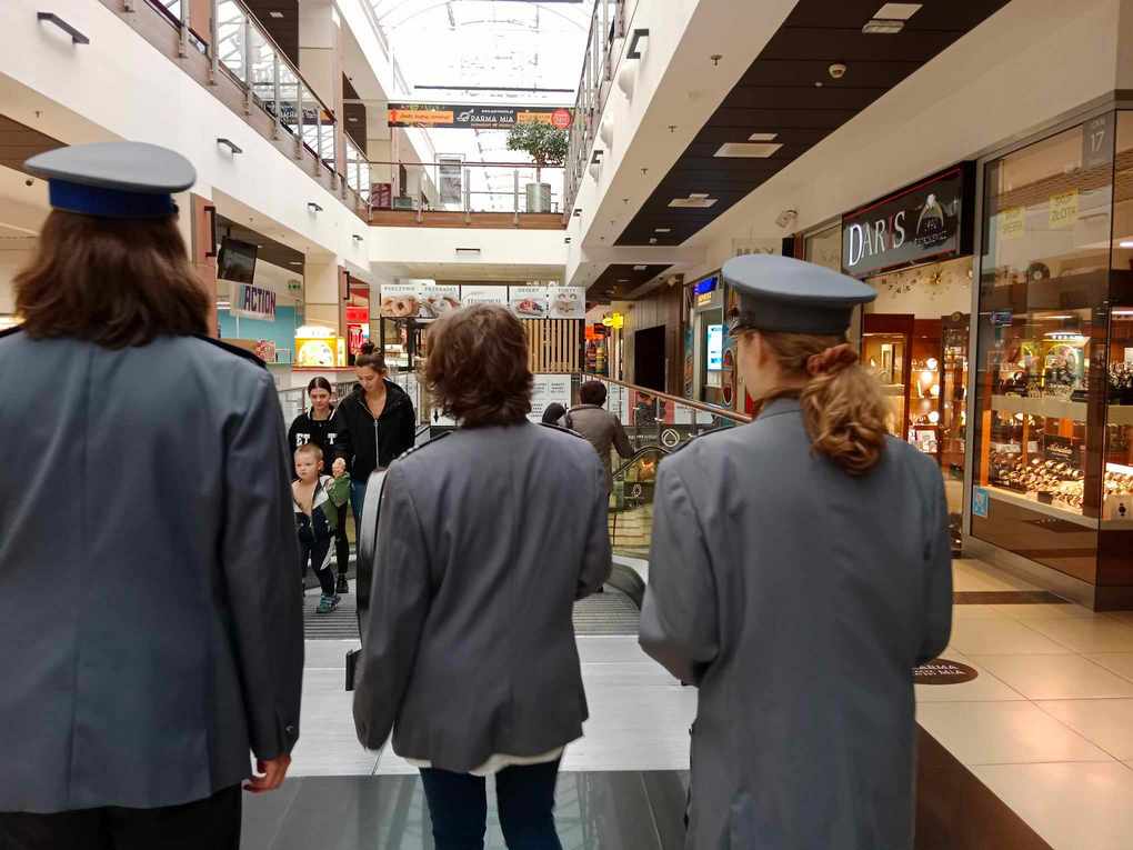 Happening policja_9 Na froncie trzy osoby ubrane w granatowe mundury policyjne. Stoją odwrócone plecami, zwrócone twarzą w stronę przechadzających się po galerii handlowej ludzi.