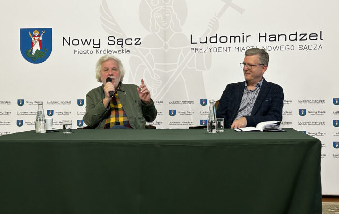 Przy dużym stole nakrytym zielonym materiałem siedzi dwóch mężczyzn. Jeden z nich przemawia do mikrofony, drugi, zwrócony w jego stronę, uśmiecha się. W tle ścianka reklamowa w jasnym kolorze.