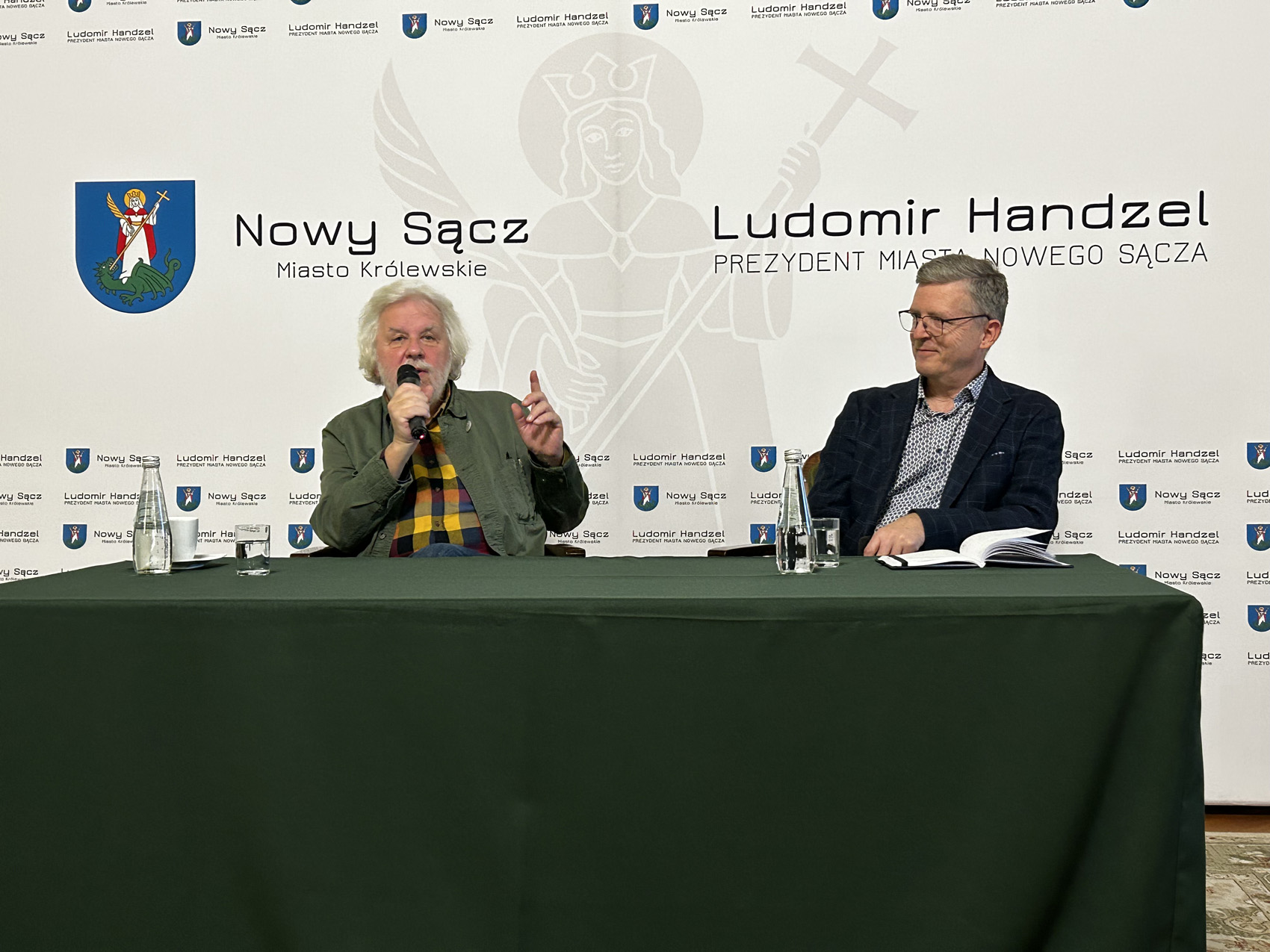 Przy dużym stole nakrytym zielonym materiałem siedzi dwóch mężczyzn. Jeden z nich przemawia do mikrofony, drugi, zwrócony w jego stronę, uśmiecha się. W tle ścianka reklamowa w jasnym kolorze.