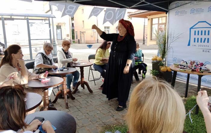 Duży namiot polowy. W centralnym miejscu ubrana w długą granatową sukienkę, czerwone korale i kolorową ludową chustkę na głowie kobieta trzyma małą laleczkę w ręku. Wokół kobiety, przy okrągłych stolikach siedzą kobiety trzymające w rękach kolorowe skrawki materiałów.