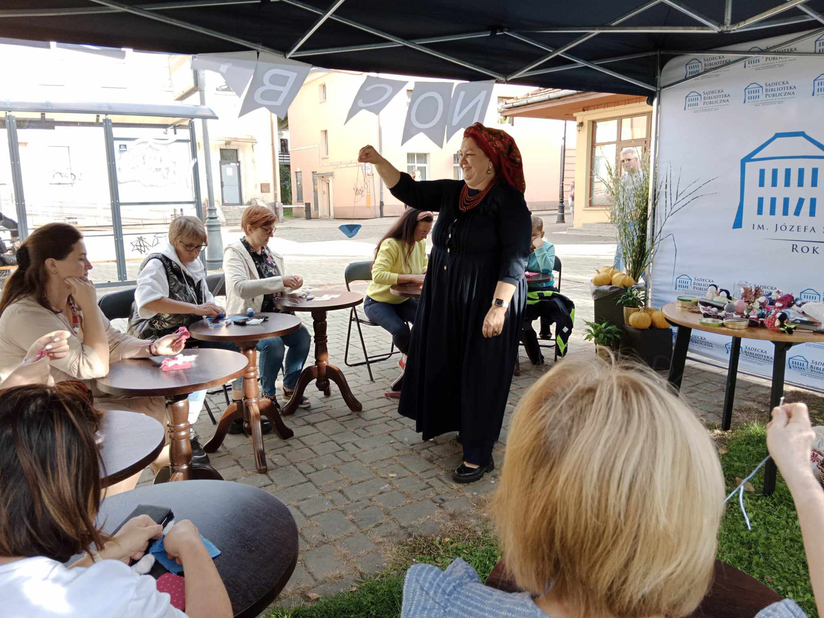 Duży namiot polowy. W centralnym miejscu ubrana w długą granatową sukienkę, czerwone korale i kolorową ludową chustkę na głowie kobieta trzyma małą laleczkę w ręku. Wokół kobiety, przy okrągłych stolikach siedzą kobiety trzymające w rękach kolorowe skrawki materiałów.