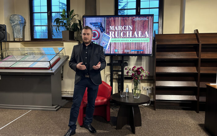 Elegancko ubrany mężczyzna stoi na środku sali. Obok niego mały brązowy stolik i czerwony fotel. Na stoliku flakon z bordowymi goździkami. W tle telewizor z kolorową grafiką.