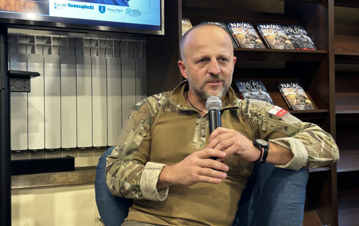 W pomieszczeniu na fotelu siedzi mężczyzna, ubrany w wojskowy strój. Mężczyzna pokazany jest od pasa w górę. W rękach trzyma mikrofon blisko ust, mówi do niego. Za mężczyzną po prawej stronie stoi długi regał z książkami.