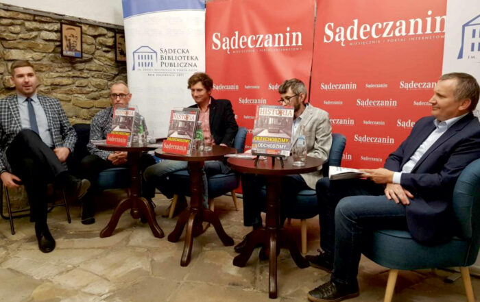 W pomieszczeniu znajduje się 5 mężczyzn. Mężczyźni siedzą w półkolu na fotelach, przed nimi stoją trzy małe okrągłe stoliki. Na stolikach stoją: trzy czasopisma, butelki z wodą, szklanki. W tle za siedzącymi rollupy: Sądeckiej Biblioteki Publicznej i Sądeczanina.