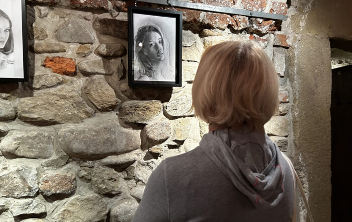 W pomieszczeniu stoi kobieta, pokazana od pasa w górę, od tyłu. Na ścianie wiszą dwa rysunki, oprawione w ramki. Kobieta patrzy na jeden z rysunków.