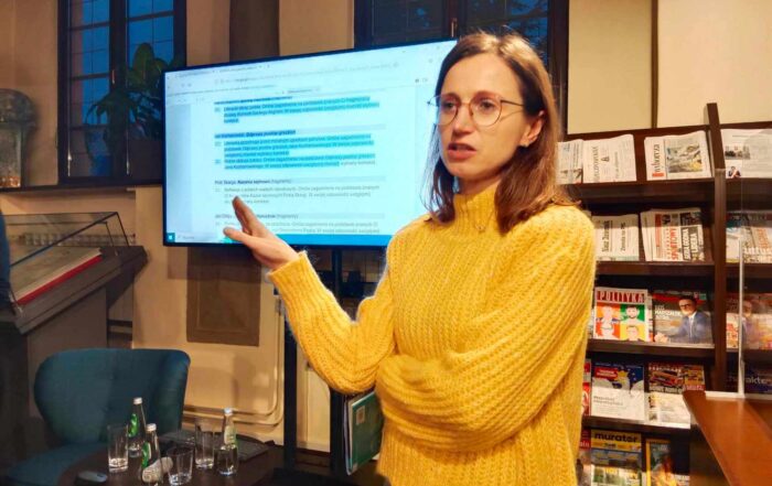 Kobieta w okularach i żółtym półgolfie wskazuje prawą ręką w stronę znajdującego się za nią telewizora. Na telewizorze wyświetla się tekst z zaznaczonymi na niebieski kolor linijkami.