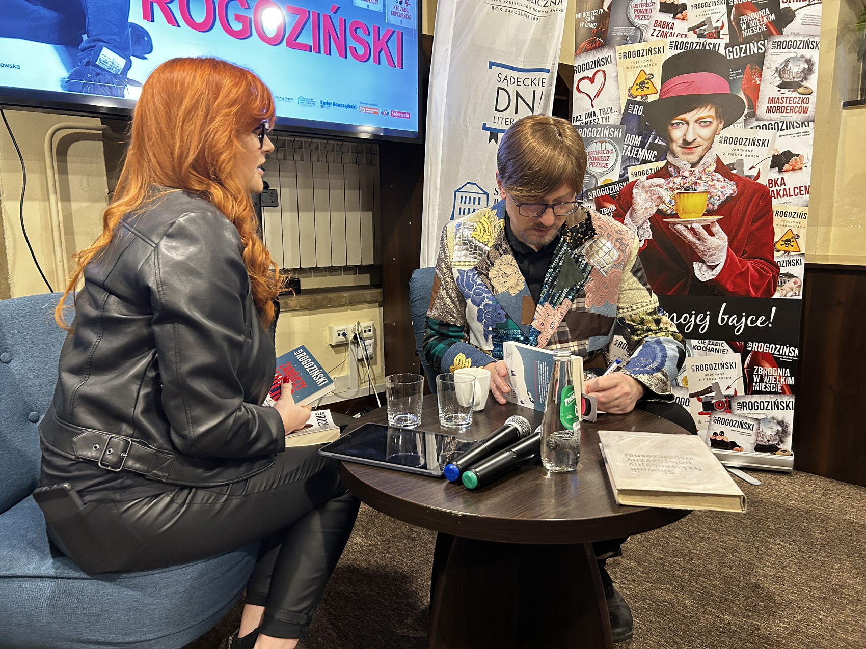 Alek Rogoziński (10) Po lewej stronie kobieta w czarnym stroju siedzi na niebieskim fotelu. Mężczyzna w kolorowej marynarce na czarnym stoliku podpisuje książkę. Za nimi znajduje się kolorowy plakat.