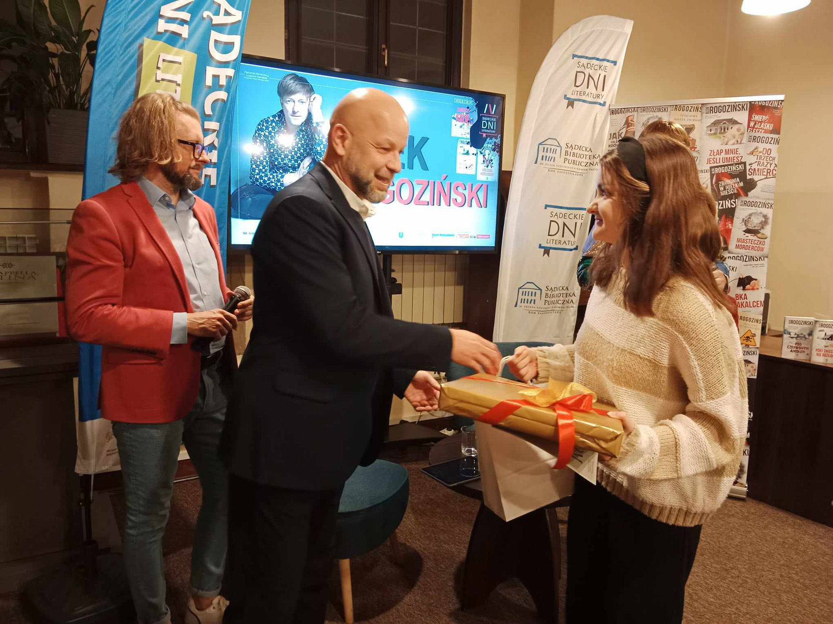 Alek Rogoziński (3) Mężczyzna w garniturze wręcza kobiecie prezent z czerwoną kokardą. Za nimi stoi mężczyzna w czerwonej marynarce i jeansach. Trzyma mikrofon. Na ścianie jest telewizor na którym wyświetlana jest sylwetka siedzącego mężczyzny. Po prawej stronie kolorowy rollup.