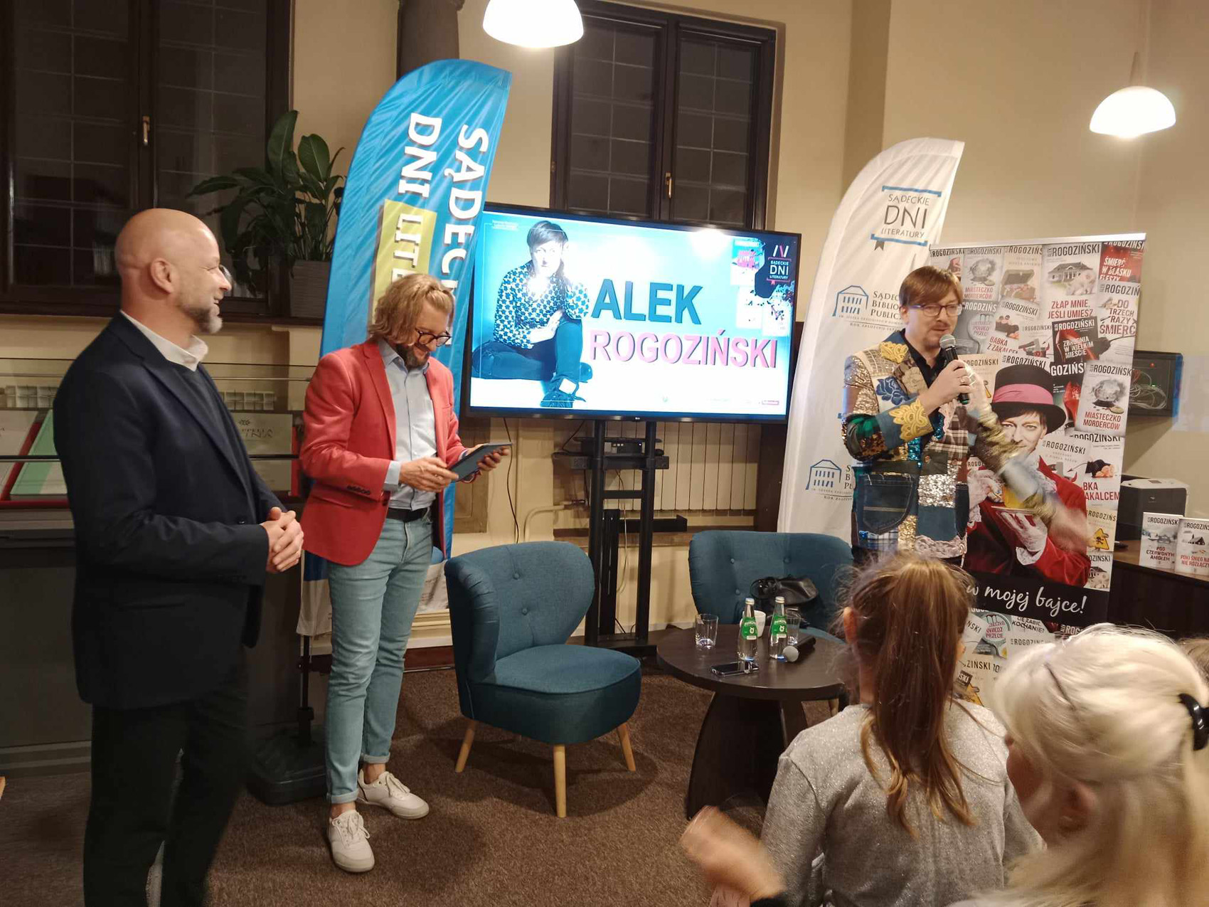 Alek Rogoziński (4) Trzech mężczyzn stoi obok foteli w kolorze niebieskim oraz stolika w kolorze czarnym. Na stoliku woda mineralna. Jeden z mężczyzn przemawia przez mikrofon. Za nimi znajduje się telewizor na którym wyświetlany jest napis Alek Rogoziński oraz sylwetka siedzącego mężczyzny Po prawej stronie stoi reklamowy rollup. W prawym dolnym rogu znajdują się dwie kobiety siedzące tyłem do fotografującego.