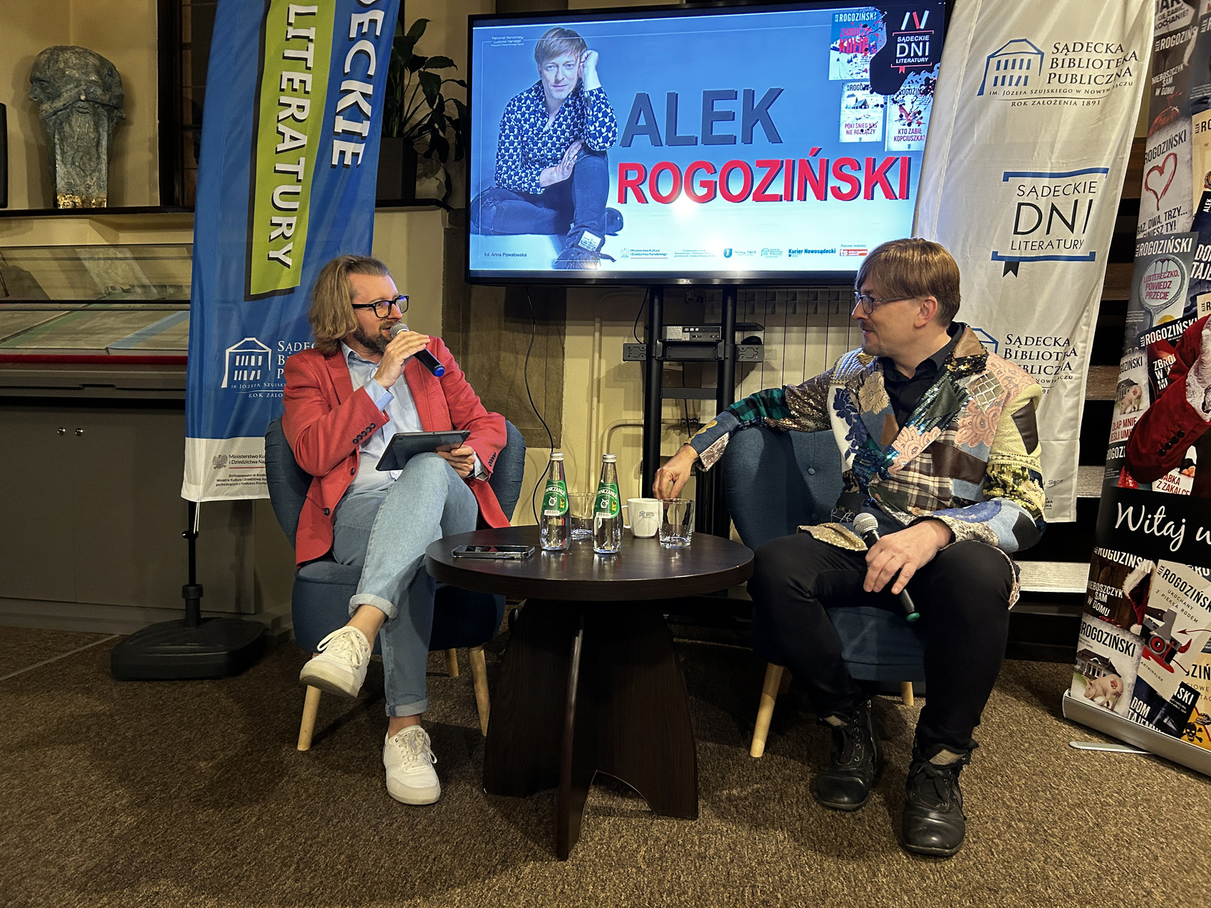 Alek Rogoziński (6) Mężczyzna w czerwonej marynarce i jeansach siedzi na niebieskim fotelu przemawia przez mikrofon. po prawej stronie siedzi mężczyzna w kolorowej marynarce i czarnych spodniach. Za nimi znajduje się telewizor na którym wyświetlany jest napis Alek Rogoziński oraz sylwetka siedzącego mężczyzny. Na środku znajduje się czarny stolik.