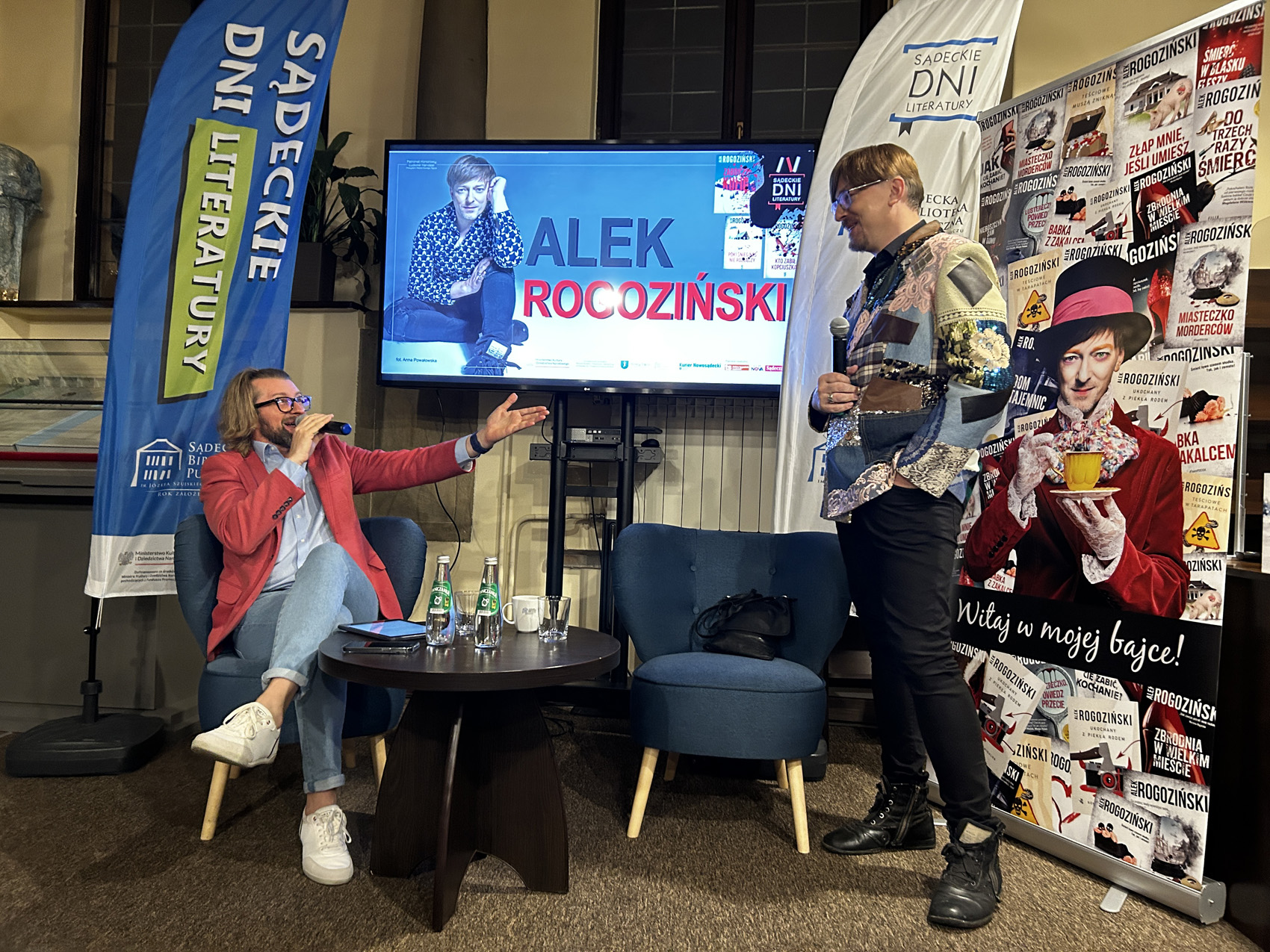 Alek Rogoziński (9) Mężczyzna w czerwonej marynarce i jeansach siedzi na niebieskim fotelu przemawia przez mikrofon. po prawej stronie stoi mężczyzna w kolorowej marynarce i czarnych spodniach. Za nimi znajduje się telewizor na którym wyświetlany jest napis Alek Rogoziński oraz sylwetka siedzącego mężczyzny. Na środku znajduje się czarny stolik. Po prawej stronie stoi kolorowy plakat.