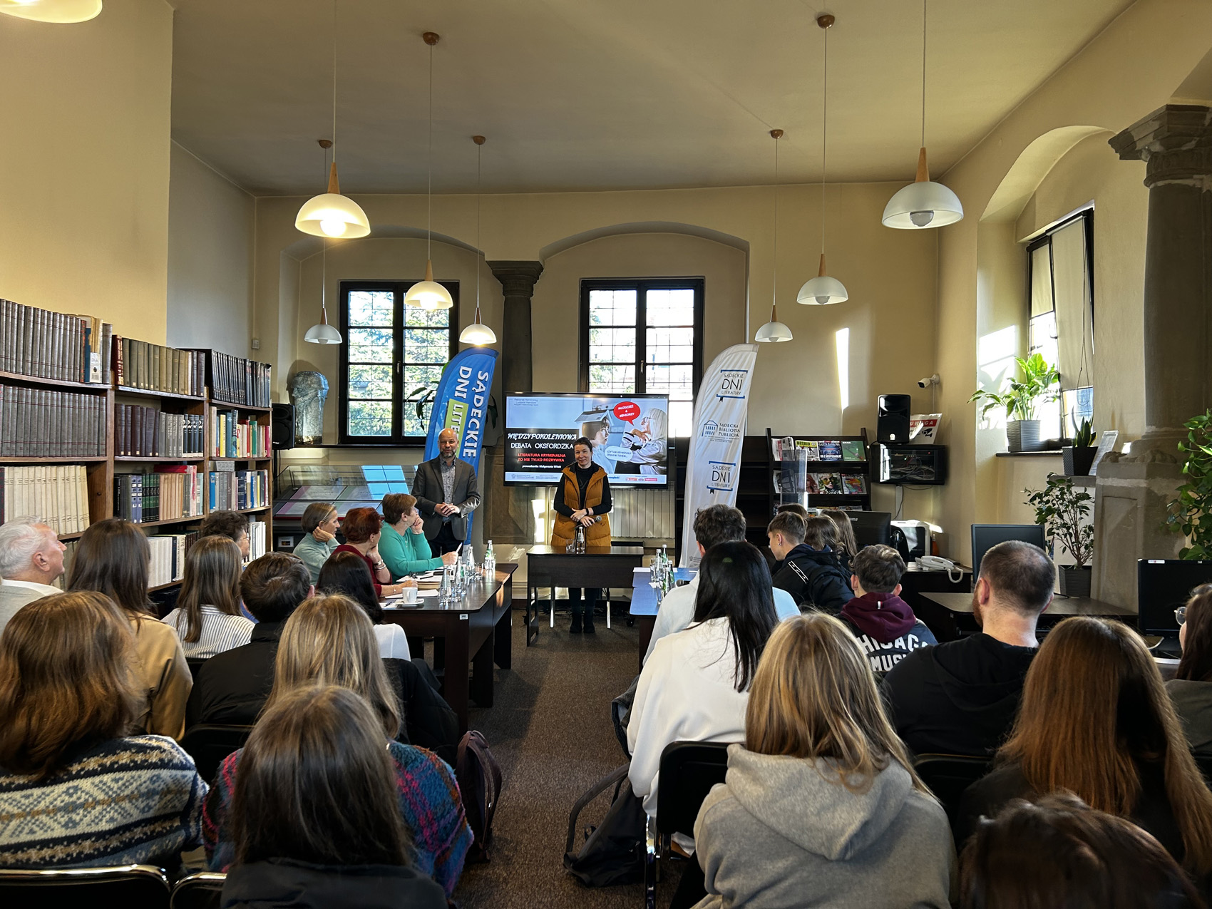 Kobieta ubrana na żółto stoi w pomieszczeniu. Przed nią brązowy stół. Na stole butelka z wodą i szklanka. Po lewej stronie niebieski baner z napisem Sądeckie Dni literatury i regał z książkami. Po prawej stronie biały baner z logotypem Sądeckiej Biblioteki Publicznej. Za kobietą na stojaku telewizor. Na telewizorze wyświetlany tekst Międzypokoleniowa Debata Oksfordzka Literatura kryminalna to nie tylko rozrywka. Grupa ludzi siedzi na krzesłach tyłem do fotografującego.