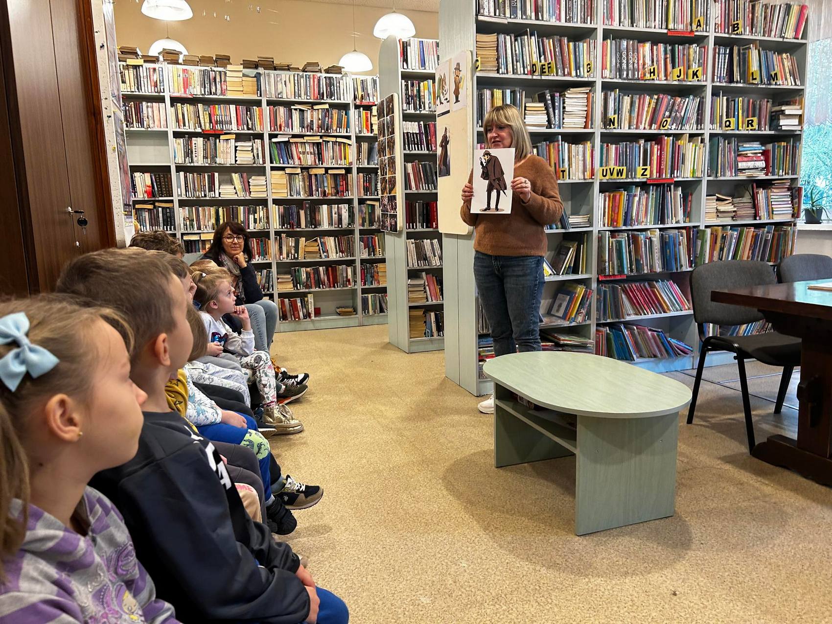 Detektyw Łodyga_1 Pokój wypełniony regałami z książkami. Po lewej stronie na ławce siedzi grupka dzieci. Przed nimi kobieta w brązowym sweterku. Pokazuje dzieciom obrazek z namalowanym detektywem.