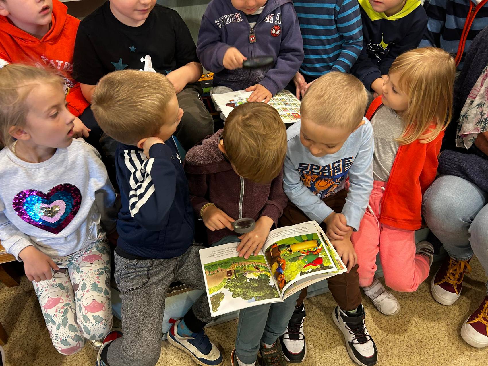 Detektyw Łodyga_2 Grupa dzieci siedzi na ławkach. Dwójka z nich trzyma na kolanach otwarte książki, na które spoglądają przez lupy.