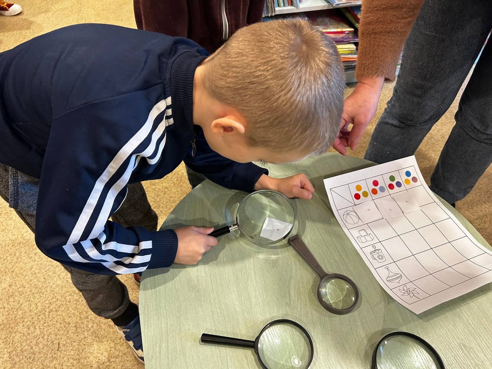 Detektyw Łodyga_4 Nad zielonym stolikiem pochyla się chłopiec. Chłopiec trzyma lupę, patrzy przez nią na karteczkę. Na stole leży papierowa plansza oraz trzy lupy.