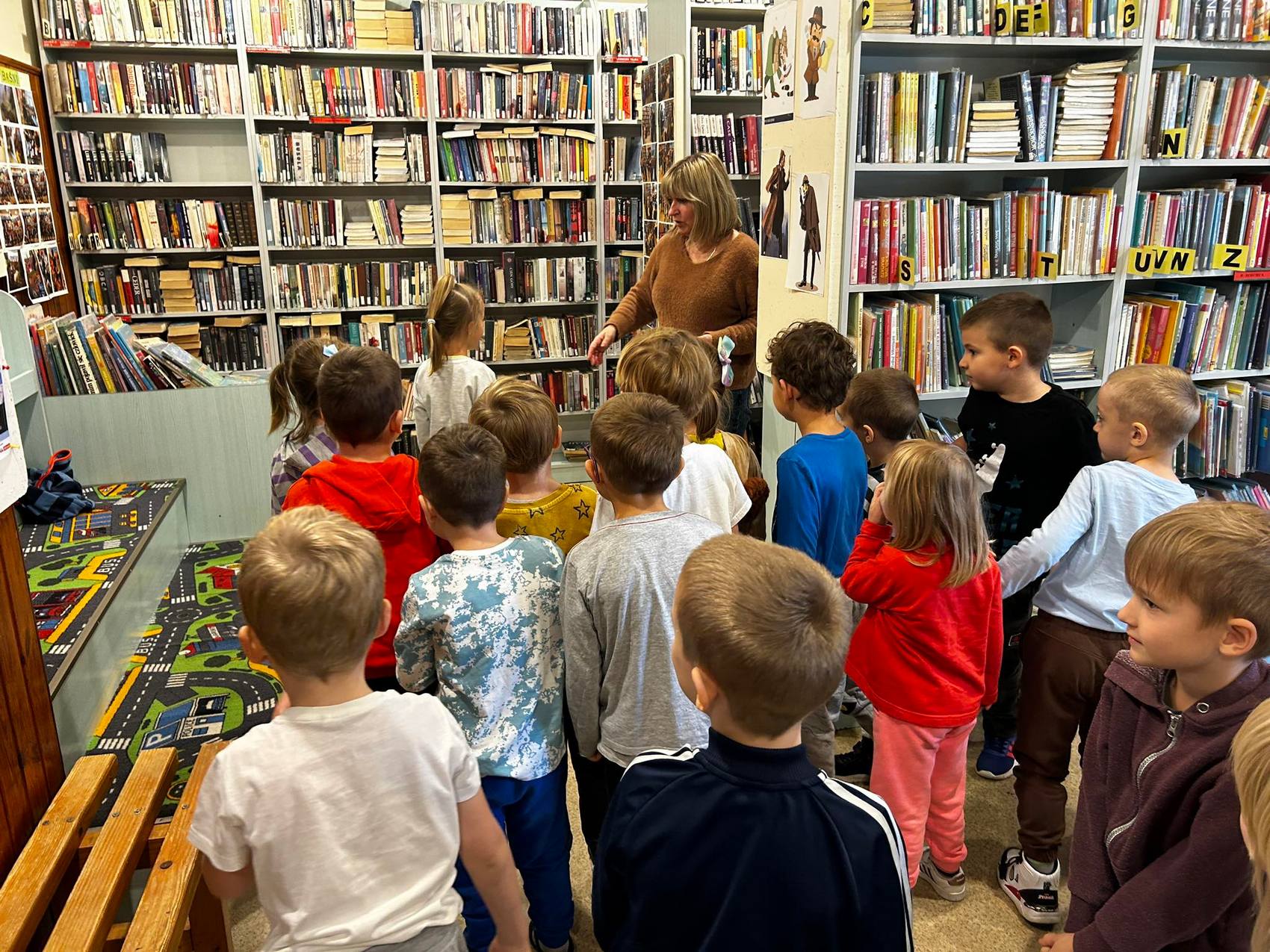 Detektyw Łodyga_6 Pokój wypełniony regałami z książkami. Na środku grupka kilkunastu dzieci zwróconych w stronę kobiety.