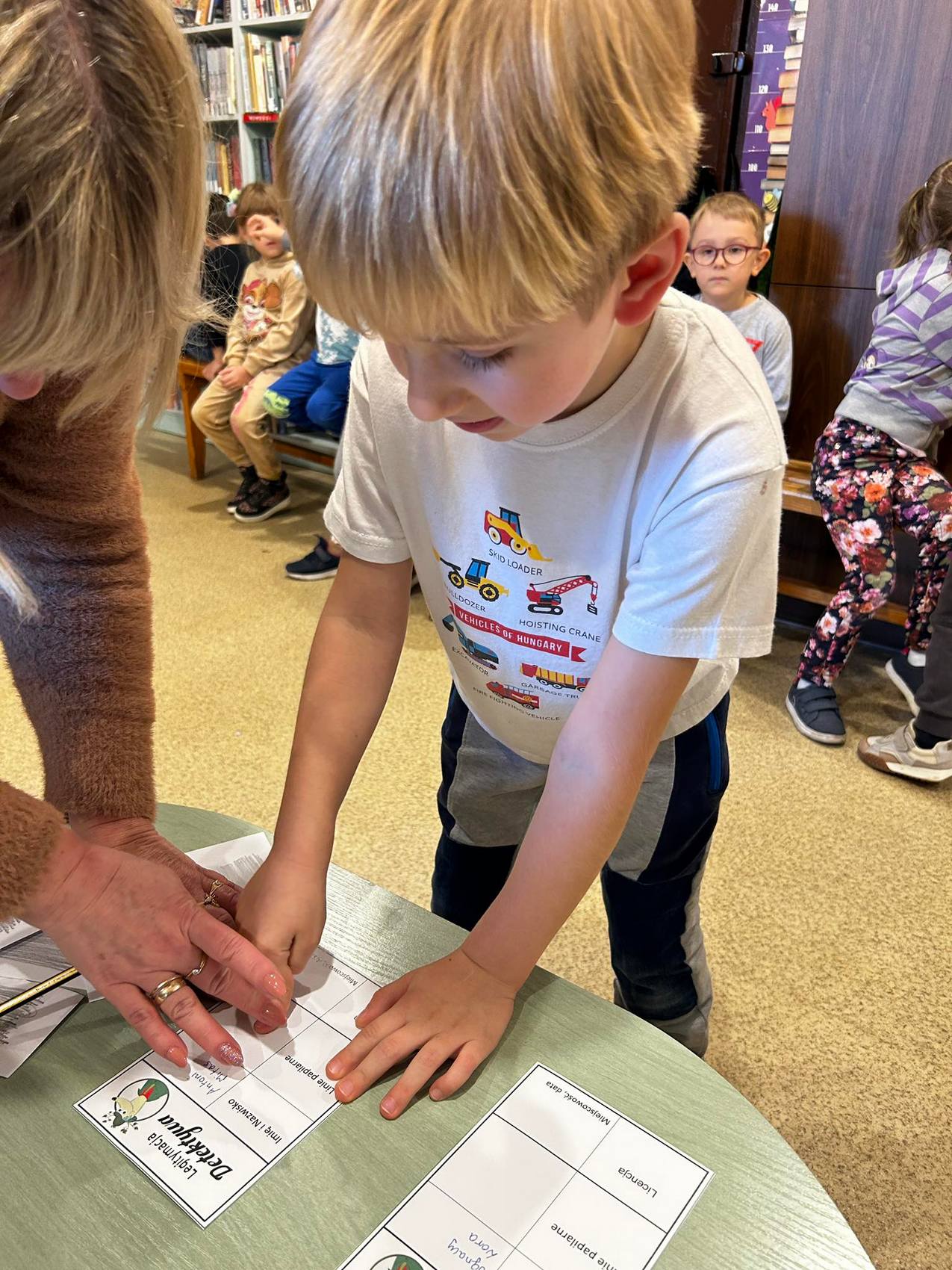 Detektyw Łodyga_7 Przy stoliku pochyla się chłopiec z kobietą. Przyglądają się rozłożonym na stole kartkom. W tle siedzące na ławce dzieci.