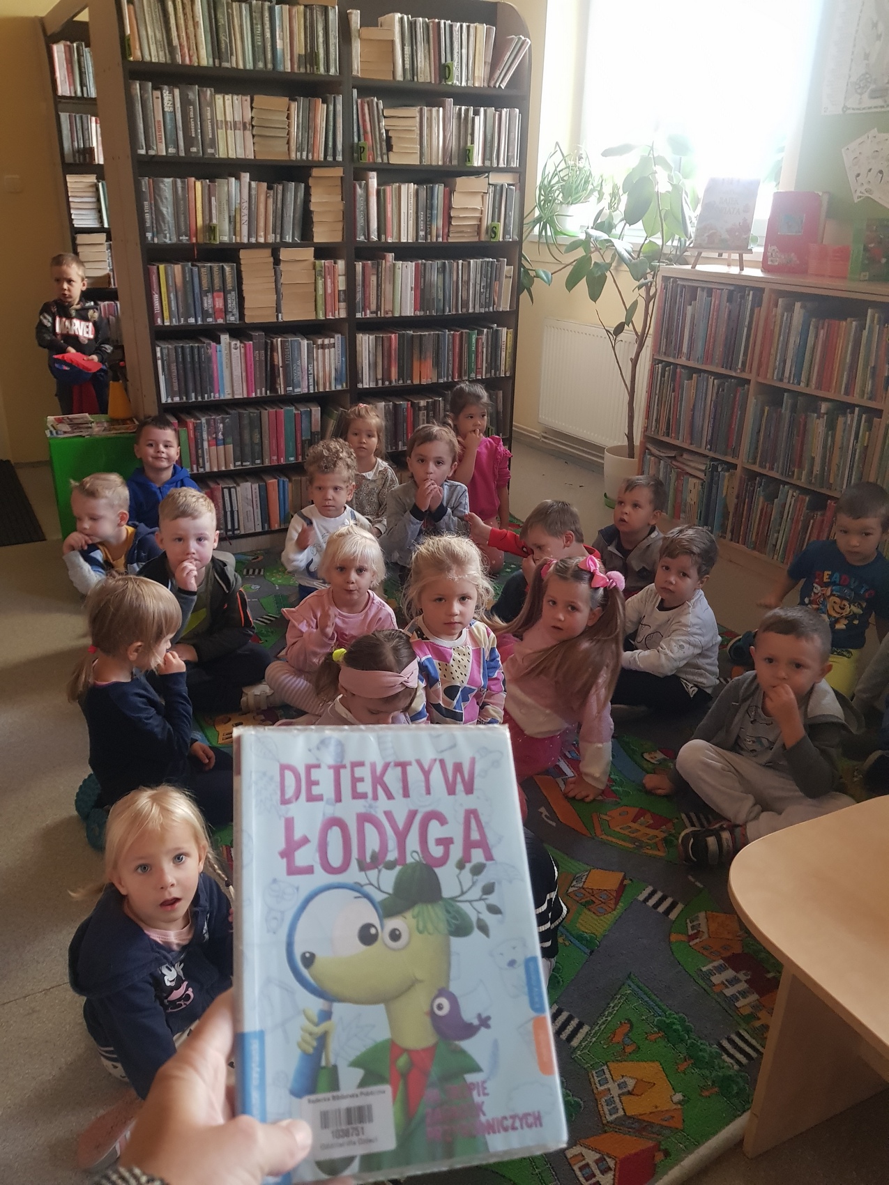 Detektyw Łodyga_8 Pokój z regałami zapełnionymi książkami. Na środku, na kolorowej wykładzinie, siedzi grupa dzieci. Na pierwszym planie fotografii dłoń trzymająca książkę.