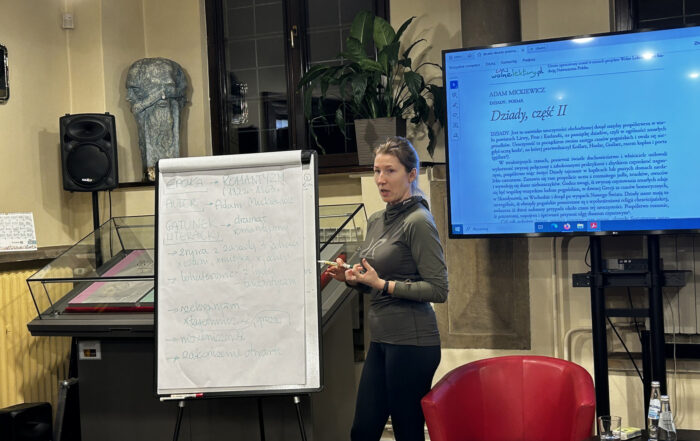 W pomieszczeniu znajduje się kobieta. Kobieta stoi na środku, trzyma w ręce pisak. Obok niej stoi flip chart. Po prawej stronie stoi czerwony fotel i mały okrągły stolik. Za fotelem stoi duży ekran.