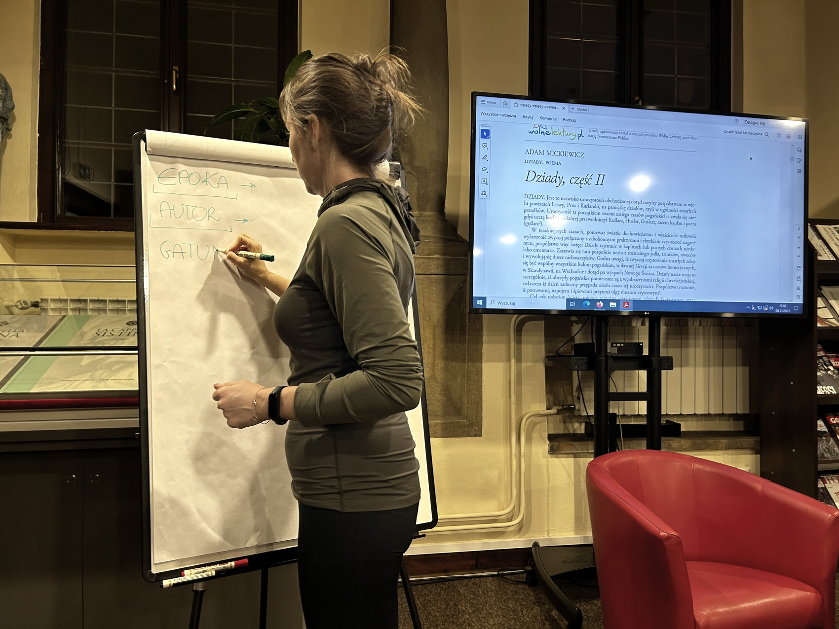 Egzamin 8klasisty – dziady ii (4) W pomieszczeniu znajduje się kobieta. Kobieta jest pokazana od tyłu. W ręce trzyma pisak. Przed nią stoi flip chart z biała kartką. Kobieta pisze pisakiem na kartce. Po prawej stronie stoi czerwony fotel. Za fotelem stoi duży ekran.