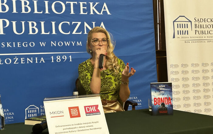 Kobieta w okularach w zielonej bluzce siedzi przy zielonym stole. Trzyma mikrofon. Na stole książka i plakat informacyjny. Z tyłu niebieski baner z logotypem Sądeckiej Biblioteki Publicznej.