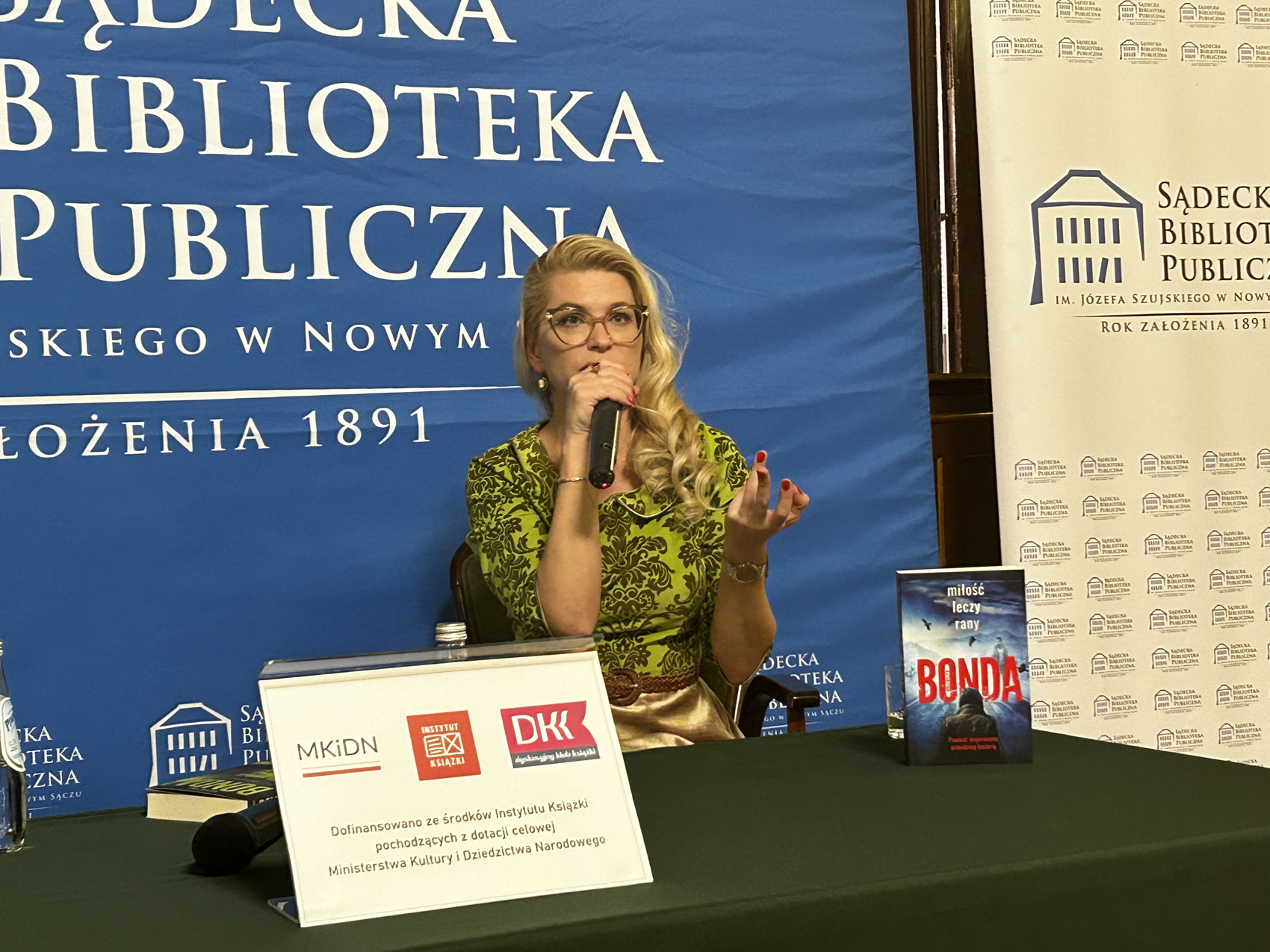 Kobieta w okularach w zielonej bluzce siedzi przy zielonym stole. Trzyma mikrofon. Na stole książka i plakat informacyjny. Z tyłu niebieski baner z logotypem Sądeckiej Biblioteki Publicznej.